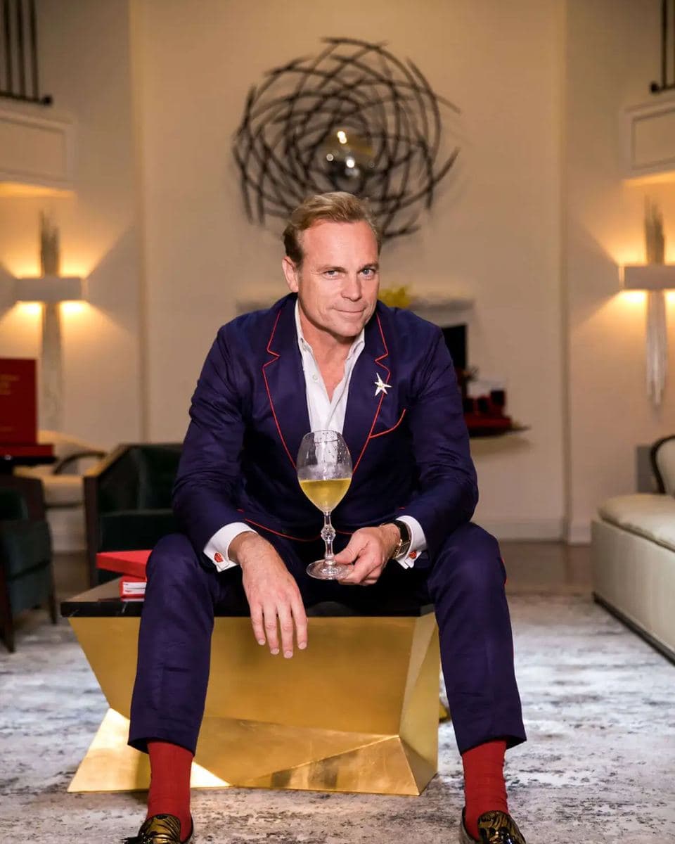 Jean-Charles Boisset, seated on a gold faceted table with a glass of wine, navy suit with red piping, red socks, gold loafers